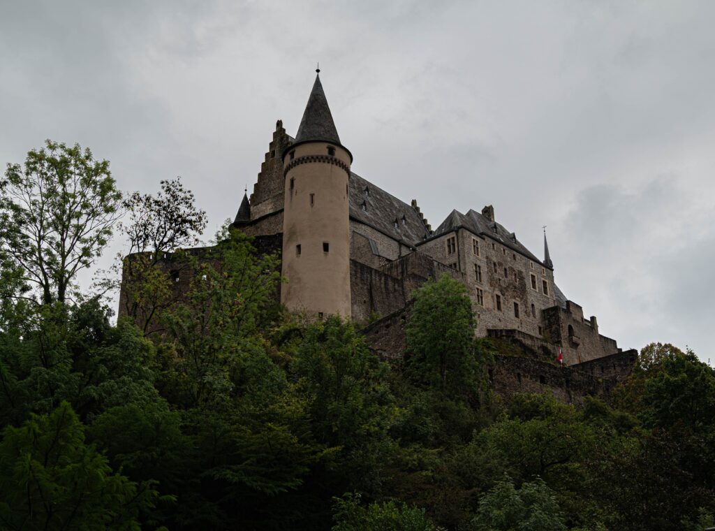 Lase vabaks see, mida sa enam ei valiks. A striking view of Vianden Castle in Luxembourg, nestled among lush greenery under a cloudy sky.