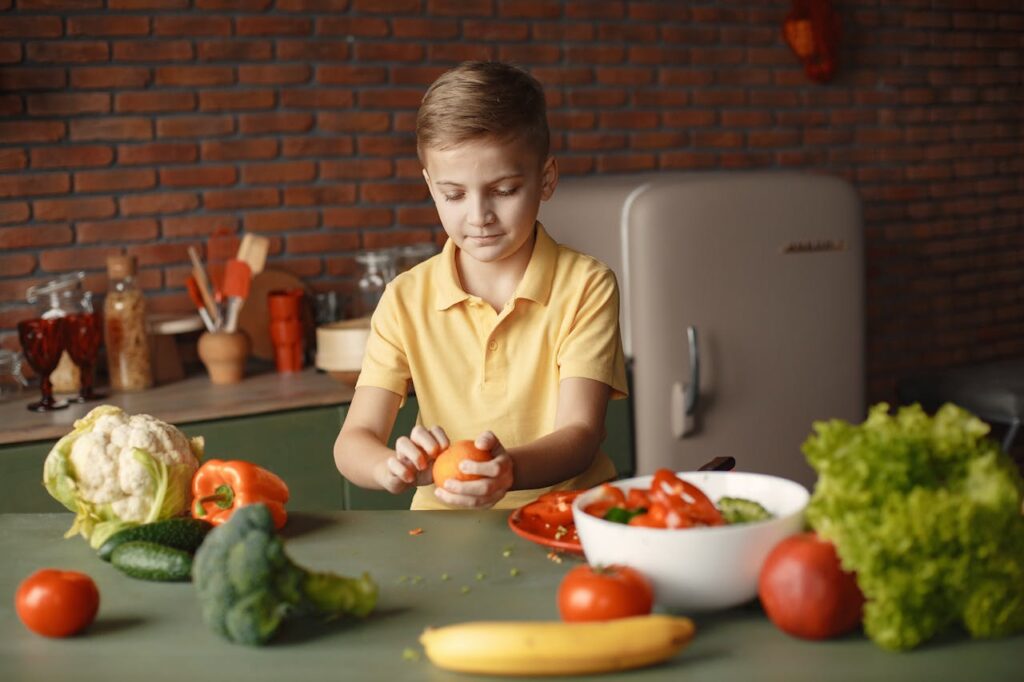 Tervislikumad harjumused erivajadustega inimestele Confident kid peeling fresh orange sitting at table with assorted vegetables and fruits near salad bowl during vegetarian meal preparation in loft kitchen with brick walls