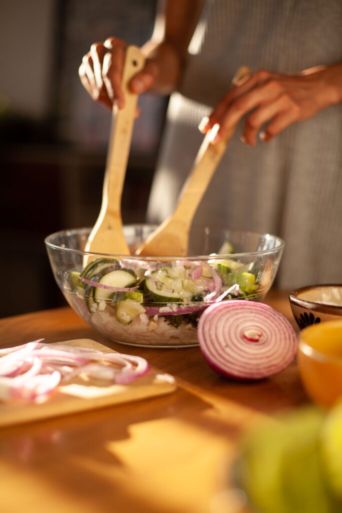 Tervislik toitumine, töökorraldus ja eeskuju kogukonna teenusel A fresh zucchini and onion salad being mixed in a glass bowl with wooden spoons indoors.