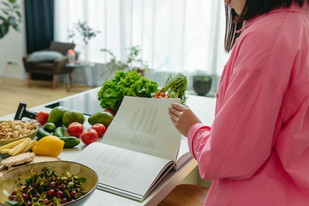 Toiduvalmistamine kui osa laagrikogemusest ja eluliste oskuste õppest A woman in a kitchen flipping through a cookbook surrounded by fresh vegetables.