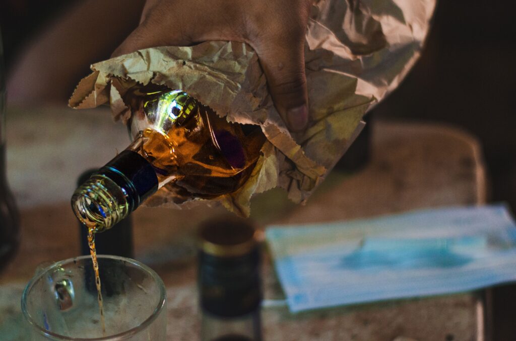 “Nonsenss Romanss” – kui töökeskkonna piirid muutuvad häguseks Close-up of a hand pouring whiskey from a paper-wrapped bottle into a glass, indoors.