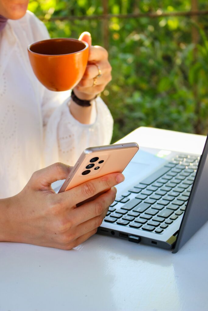 Mõnikord just nii paranemine välja näebki. Person using a smartphone while drinking coffee near a laptop outdoors, showcasing multitasking with technology.