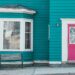 Exterior of a teal house featuring a red door, window, and bench on a sunny day.